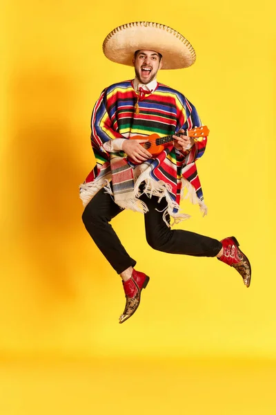 カラフルなポンチョを着た若い感情的な男とポーズ ギターを演奏し 黄色のスタジオの背景にジャンプします メキシコの伝統 楽しさ お祝い 感情の概念 — ストック写真