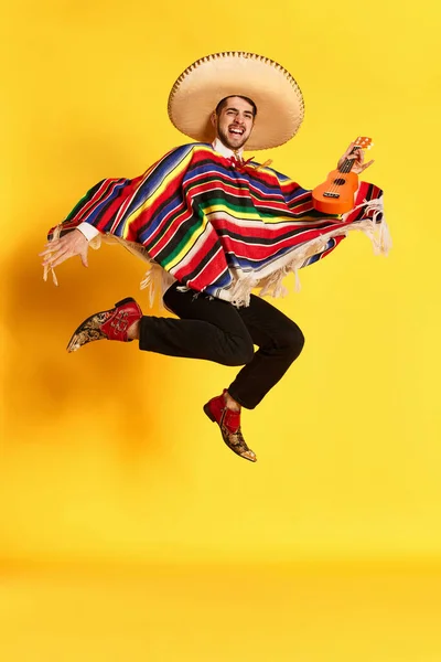 カラフルなお祝いの服を着た若い興奮した男 ポンチョと黄色のスタジオの背景にギターでジャンプするポーズのいくつかのブレロ メキシコの伝統 楽しさ お祝い 感情の概念 — ストック写真
