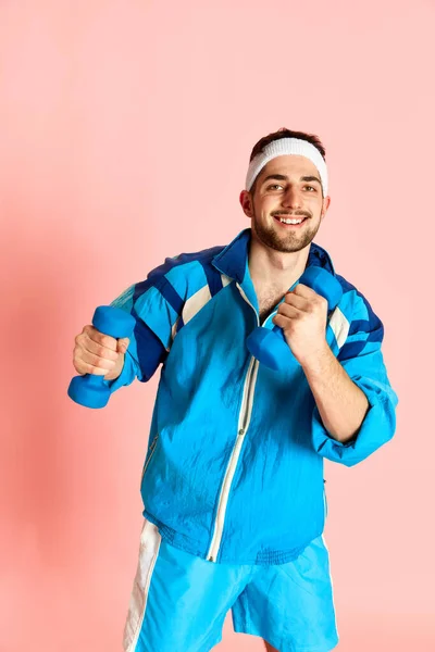 Portrait of young active sportive man in blue sportswear training, posing with dumbbells against pink studio background. Concept of sportive lifestyle, emotions, facial expression. Ad