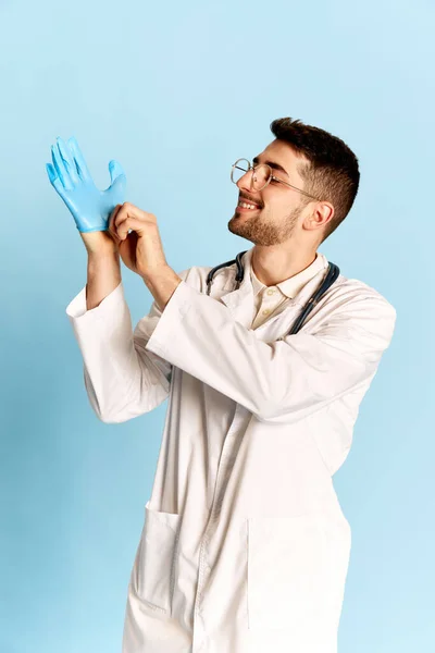 若い正のひげを生やした男の肖像画 白い医療用ガウンの医師は ゴム手袋をはめて 青のスタジオの背景に対してポーズ 職業の概念 — ストック写真