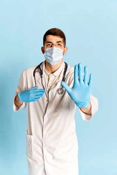 青のスタジオの背景に対してポーズを若い男 白い医療ガウン 保護マスクや手袋の医師の肖像画 病気の予防 職業の概念 — ストック写真