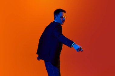 Çok hoş görünüyor. Genç neşeli bir adamın portresi, ceketli bir öğrenci, neon ışıkta turuncu stüdyonun arka planında dans ediyor. Gençlik kavramı, duygular, yüz ifadesi, yaşam tarzı. Ad