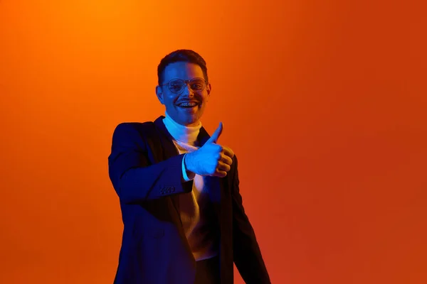 stock image Portrait of delightful happy young man in jacket and glasses posing with positive finger gesture against orange studio background in neon light. Youth, emotions, facial expression, lifestyle concept