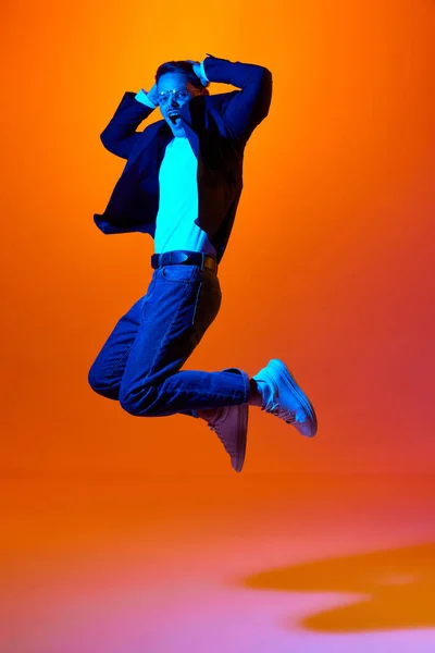 stock image Full-length portrait of young emotive man in stylish casual clothes emotinally jumping against orange studio background in neon light.. Concept of youth, emotions, facial expression, lifestyle. Ad