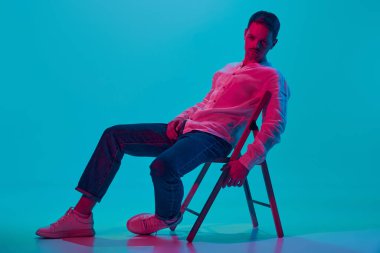 Ciddi bir bakış. Beyaz gömlekli ve kot pantolonlu genç adam portresi sandalyede oturuyor, pembe neon ışıklı mavi stüdyo arka planına karşı poz veriyor. Gençlik kavramı, duygular, yüz ifadesi, yaşam tarzı. Ad
