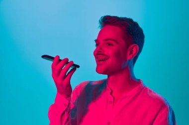 Gülümseyen mutlu adamın portresi cep telefonuyla pembe neon ışıkta mavi stüdyonun arka planına sesli mesaj kaydediyor. Gençlik kavramı, duygular, yüz ifadesi, yaşam tarzı. Ad