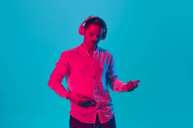 Beyaz gömlekli genç bir adamın portresi kulaklıkla müzik dinlerken ve illüzyon gitarıyla pembe neon ışıkta mavi stüdyo arka planına karşı çalarken. Gençlik kavramı, duygular, yüz ifadesi.