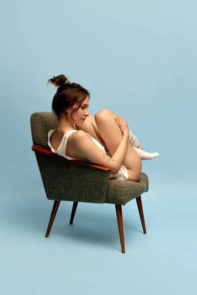 stock image Feeling comfortable and cozy. Portrait of young slim girl with hairbun, in home wear and socks posing in armchair against blue studio background. Concept of body and skin care, figure, wellness.