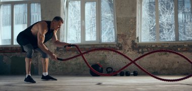 Konsantre genç adam sportif, kaslı, güçlü vücut eğitimli ve kapalı alanda savaş halatları kullanıyor. Antrenman yapıyorum. Spor yaşam tarzı, vücut bakımı, fitness, hobi, sağlık, eylem ve hareket kavramı. Pankart