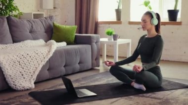 Spor giyimli, sakin, genç bir kız kulaklıklı bir fitness adamının üzerine oturmuş online meditasyonlar dinliyor. Evde yoga eğitimi. Sportif ve sağlıklı yaşam tarzı kavramı, sağlık, bakım.