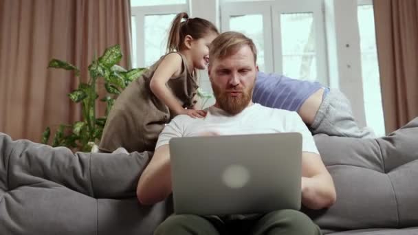 成熟的男人 父亲坐在客厅的沙发上远程工作 小孩子在他身边玩耍 分散他的注意力 自由职业 远程工作概念 — 图库视频影像