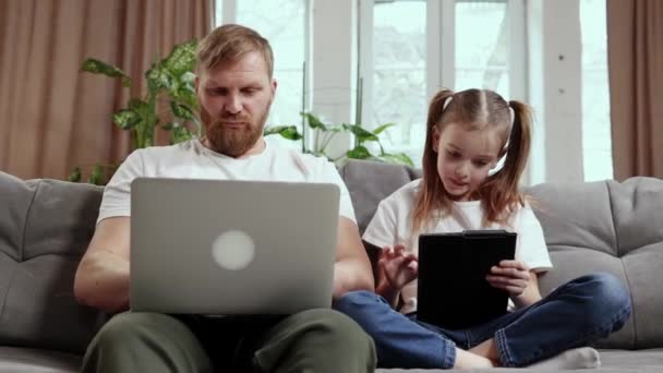 家族の時間だ 成熟したひげを生やした男は 父親が自宅のノートパソコンでリモートで作業し 小さな娘がタブレットで遊んでいるソファに座っていました 子供時代 フリーランスの仕事 リモートワークの概念 — ストック動画