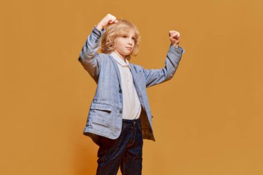 Kazandım. Başarı. Küçük bir çocuk portresi, sarı kıvırcık saçlı çocuk turuncu stüdyo arka planında resmi olarak poz veriyor. Çocukluk kavramı, duygular, yüz ifadesi, yaşam tarzı. Ad