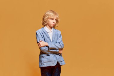 Küçük çocuk, sarı kıvırcık saçlı sakin, ciddi ve sıkılmış yüzüyle turuncu stüdyo geçmişine poz veren çocuk. Çocukluk kavramı, duygular, yüz ifadesi, yaşam tarzı. Ad