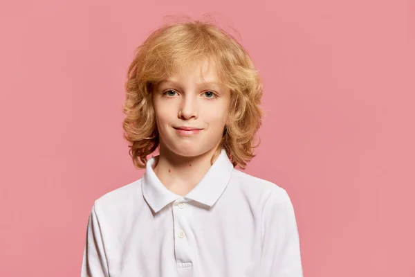 Portrait of little boy, child, pupil with curly blonde hair in white shirt looking at camera with smile against pink studio background. Concept of childhood, emotions, facial expression. Ad