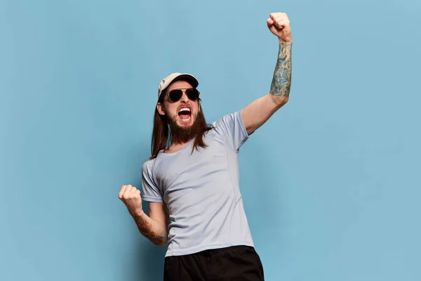 Portrait of young man with long hair posing in cap and sunglasses against blue studio background. Watching sport match. Cheering up team. Concept of emotions, facial expression, lifestyle
