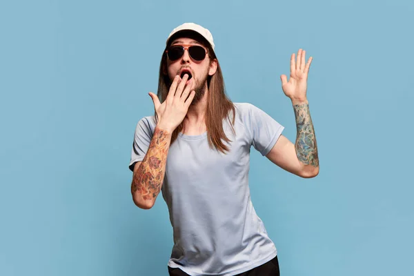 Portrait of young man with long hair posing in cap and sunglasses against blue studio background. Shocked, surprised face. Concept of emotions, facial expression, lifestyle, sport fan