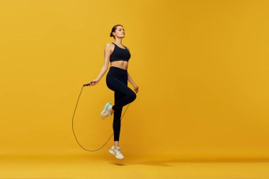 Formda vücut eğitimi almış genç bir kız sarı stüdyo geçmişiyle ip atlayarak atlıyor. Kardiyo çalışması. Spor anlayışı, sağlıklı ve aktif yaşam tarzı, güzellik, fitness