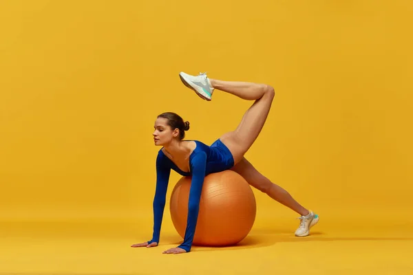 做伸展运动 漂亮的年轻姑娘穿着蓝色的运动服 带着黄色背景的健身球进行训练 健康和积极的生活方式 优雅的概念 — 图库照片