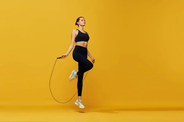 黄色のスタジオの背景にジャンプロープを飛び越える フィットボディトレーニングとスポーティな若い女の子 心臓のトレーニングだ スポーツ 健康的でアクティブなライフスタイル 美しさ フィットネスの概念 — ストック写真