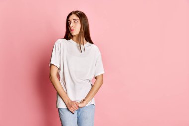 Rahat giysiler içinde genç güzel bir kızın portresi. Pembe stüdyo arka planına bakarak sakince dikiliyor. Kararsız ve utangaç hissediyorum. Gençlik kavramı, duygular, yüz ifadesi, yaşam tarzı