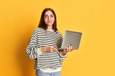 Sarı arka planda dizüstü bilgisayarla poz veren çizgili süveterli genç kız. İş fuarı ve öğrenci hayatı. Gençlik kavramı, duygular, yüz ifadesi, yaşam tarzı, eğitim, iş