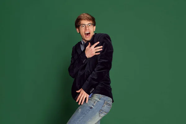 Feeling shy. Handsome young man in glasses covering body with hands with wide open mouth and eyes against green studio background. Concept of business, education, fashion, emotions, youth, lifestyle