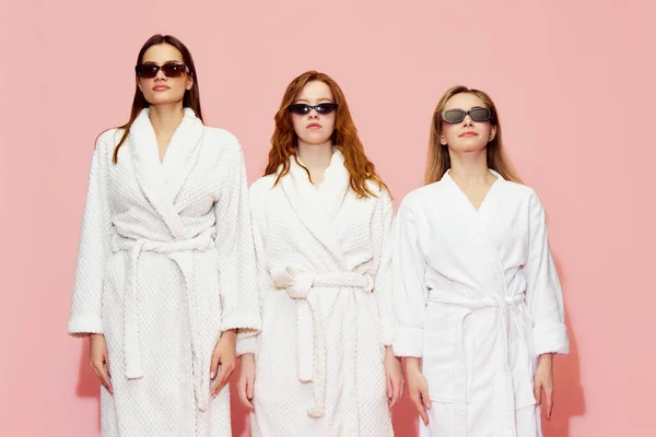 三个穿着白色浴衣和太阳镜的年轻漂亮姑娘的画像 在粉红的工作室背景下表现出严肃的表情 放松的概念 — 图库照片