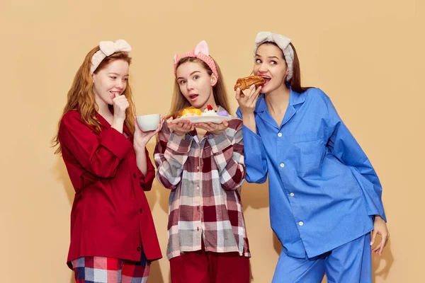 过夜的人穿着五颜六色舒适睡衣的漂亮姑娘们吃着糖果 在橙色工作室的背景下玩得很开心 放松的概念 — 图库照片