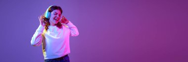 Duygusal, güzel, gündelik giysiler içinde kulaklıkla müzik dinleyen ve neon ışıkta mor arka planda şarkı söyleyen genç bir kız. Duygular, gençlik, yaşam tarzı. Pankart. İlan için alanı kopyala