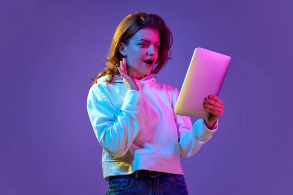 美しい若い女の子感情的にタブレットでは ネオンの光の中で紫色の背景に笑みを浮かべている オンラインコミュニケーションと良いニュース ライフスタイル フリーランスの仕事の概念 — ストック写真