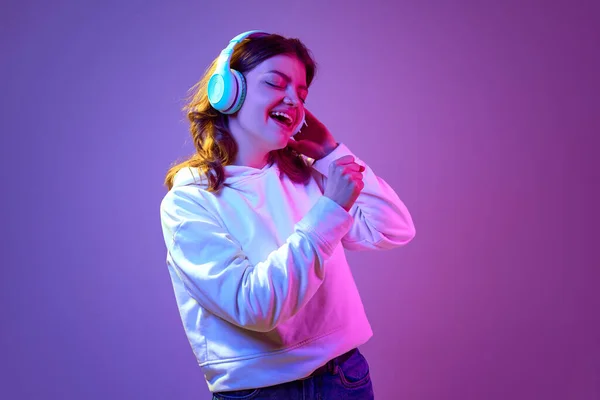 気分がいい ヘッドフォンで音楽を聴き ネオンの光の中で紫のスタジオの背景を背景に歌う幸せと肯定的な若い女の子 ライフスタイル 楽しさ ファッションの概念 — ストック写真