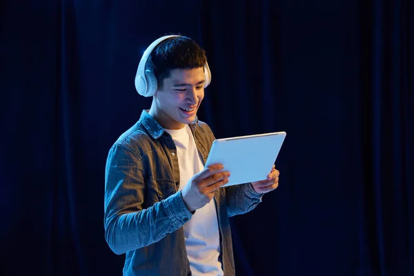Young guy playing online video games, listening to music in headphones, looking on tablet against dark background in neon light. Concept of human emotions, youth, fashion, lifestyle