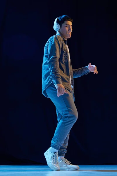 Full-length image of young guy in casual clothes listening to music in headphones and dancing against dark background in neon light. Concept of human emotions, youth, fashion, lifestyle