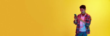 Mutlu, güler yüzlü genç adam cep telefonuna bakıyor, sarı arka planda sarı ışıkta mesajlaşıyor. Duygular, gençlik, moda, yaşam tarzı kavramı. Pankart. İlan için alanı kopyala