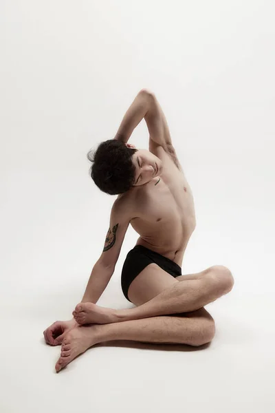 Morning stretching. Young guy sitting on floor shirtless in underwear against white studio background. Slim fit body shape. Concept of male body aesthetics, style, fashion, health, mens beauty
