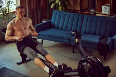 Sabit kürek makinesiyle üstsüz antrenman yapan, gündüzleri evde egzersiz yapan kaslı genç bir adam. Evde spor salonu. Spor yaşam tarzı, vücut ve sağlık, spor ve sağlık anlayışı