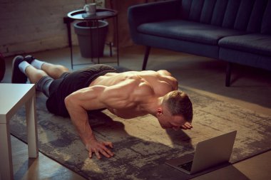 Güçlü, kaslı vücut eğitimli, spor bir genç, evde dizüstü bilgisayar dersleri alarak şınav çekiyor. Spor yaşam tarzı, vücut ve sağlık, spor ve sağlık anlayışı