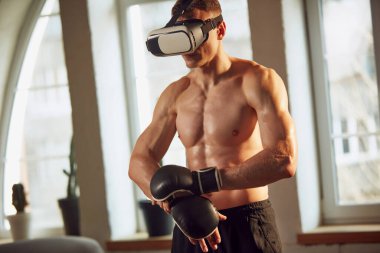 Kaslı, rahatlatıcı, güçlü, çekici vücudu olan, VR gözlük takan, evde tişörtsüz ve online dersleri olan bir sporcu. Spor yaşam tarzı, vücut ve sağlık hizmetleri, fitness, sağlık konsepti