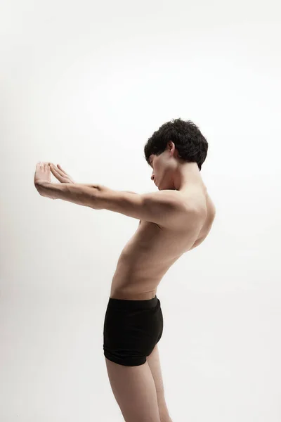 Stretching. Studio image of young guy, man standing shirtless in underwear against white background. Slim body shape. Concept of male body aesthetics, style, fashion, health, mens beauty