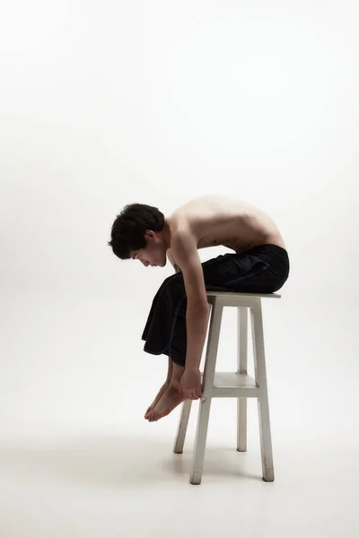 Concentration on inner self. Image of young man posing shirtless in black trousers on high chair against white studio background. Concept of male body aesthetics, style, fashion, health, mens beauty