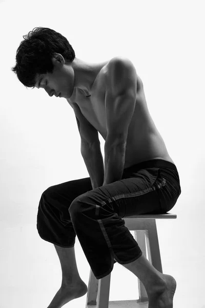 黒と白のハンサムな男のイメージが白いスタジオの背景に対して高い椅子の上に黒のズボンの中に恥ずかしそうにポーズ 男性の体の美学 スタイル ファッション メンズ美しさの概念 — ストック写真