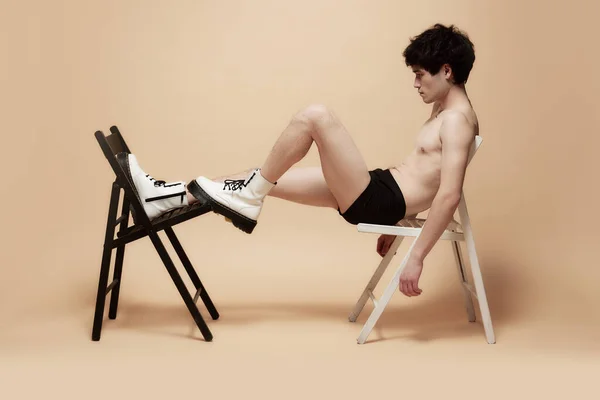 下着やスタイリッシュなブーツの若いアジアの男は ライトブラウンのスタジオの背景に対して椅子の上に軽率なポーズ 男性の体の美学 スタイル ファッション メンズ美しさの概念 — ストック写真