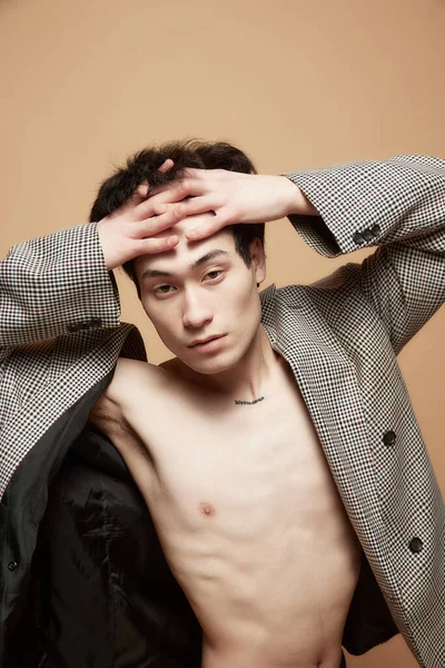 Portrait of young asian man posing shirtless in stylish grey jacket against light brown studio background. Concept of male body aesthetics, style, health, mens beauty and fashion
