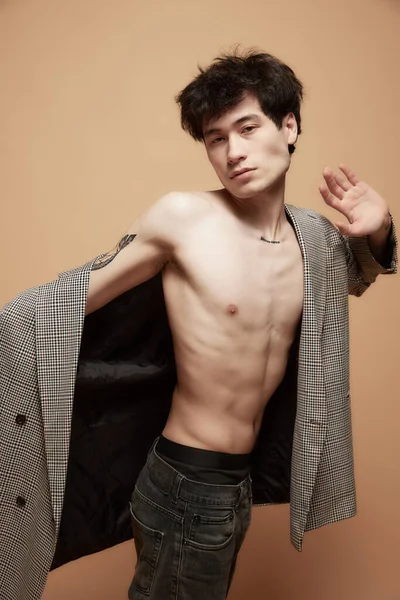 Portrait of young asian guy with relief body posing shirtless in stylish grey jacket against light brown studio background. Concept of male body aesthetics, style, health, mens beauty and fashion