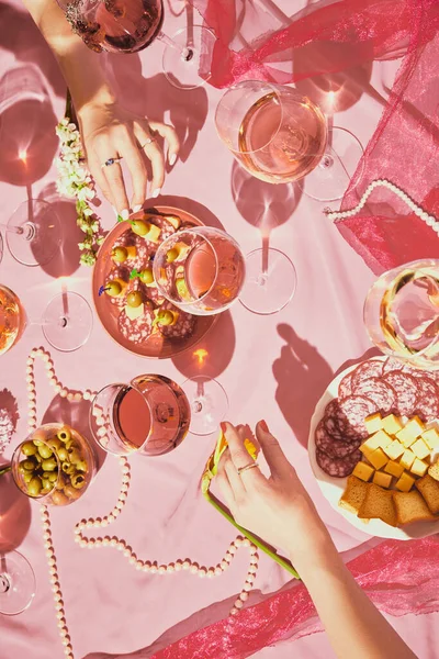 Vertical image of female hands over table with rose wine and appetizers, snacks on pink textured background, tablecloth. Concept of taste, alcohol, wine degustation, celebration, winemaking. Flat lay