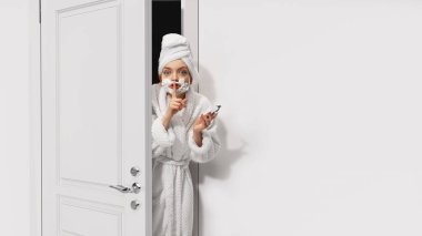 Bornozlu, havlulu ve tıraş köpüklü güzel bir kızın beyaz kapıyı dikizlediği komik bir görüntü. Sır saklamak, saklamak. Duyguların kavramı, yüz ifadesi, yaşam tarzı, gençlik, güzellik