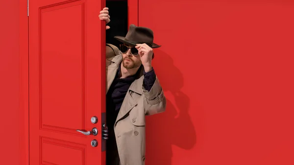 Stylowy Brodaty Mężczyzna Płaszczu Okularach Detektyw Podglądający Czerwone Drzwi Uważnie — Zdjęcie stockowe