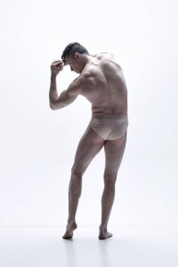 Gömleksiz, kaslı, sırt üstü duran, beyaz stüdyo arka planında iç çamaşırı pozu veren bir adamın tam boy dikiz görüntüsü. Erkek vücudu estetiği, erkek güzelliği, ilham.