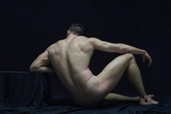 Statue. Back view portrait of muscular shirtless man sitting and posing in underwear against black studio background. Like antique sculpture. Concept of body aesthetics, mens beauty, inspiration, art